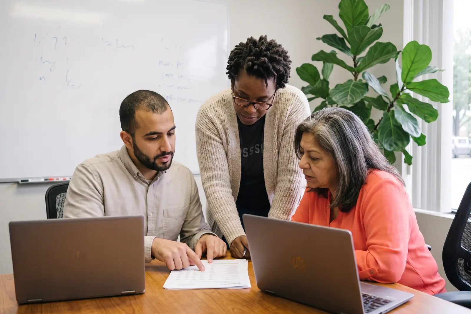 Dana, Omar and Lupe around a small conference table with laptops and printed reports. Sample photo with invented people.