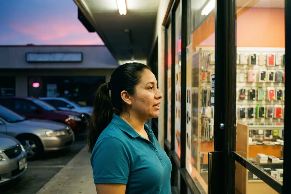 Karla at the door of the Oak Cliff store at dusk, the lit counter and phone cases behind her. Sample photo with invented people.